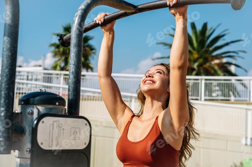 Preview: Young woman performing pull-ups on outdoor gym equipment by the beach, smiling at the sky