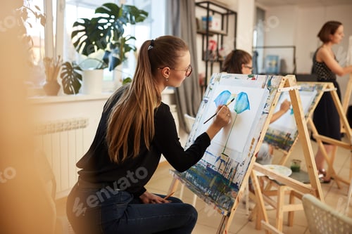 Preview: Charming fair-haired girl in glasses dressed in black blouse and jeans sits at the easel and paints