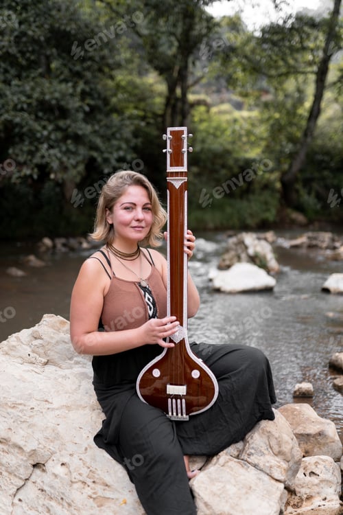 Preview: Portrait of a young adult woman posing with her indian tanpura sitting on a river rock