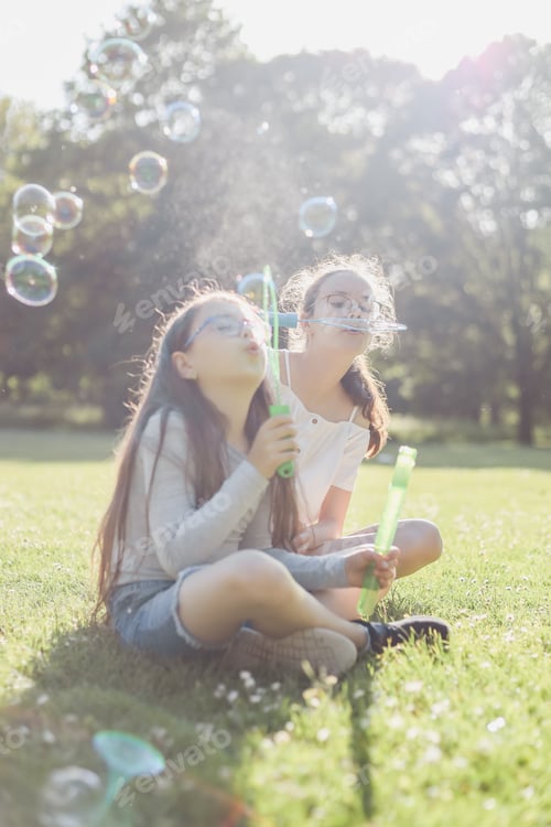 Preview: Portrait of two beautiful caucasian girl sisters blow bubbles sitting cross-legged on a flower meado