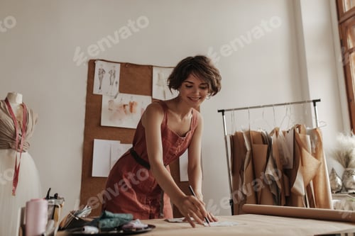 Preview: Happy cheerful short-haired woman in red linen dress smiles sincerely, works as fashion designer. B