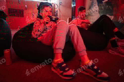 Preview: Two boys gamers play gamepad video game console in red gaming room.