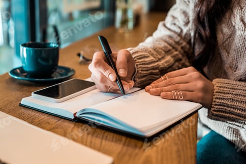 Preview: cropped view of freelancer writing in planner with smartphone in cafe