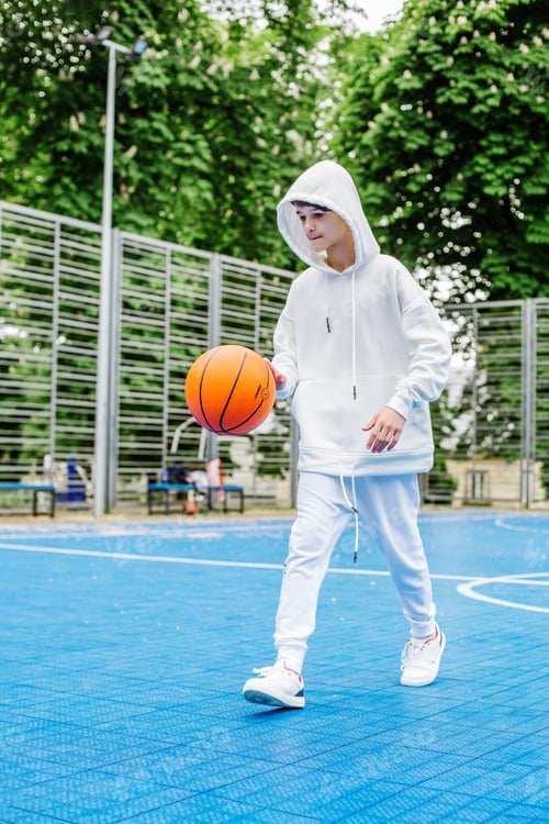 Preview: Teenage boy plays basketball on basketball court outside near school. White tracksuit.
