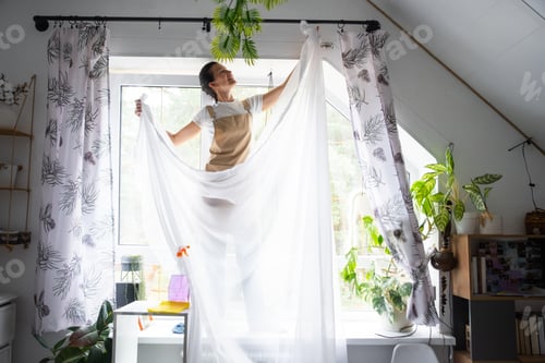 Preview: Woman in apron hangs transparent tulle curtains on large attic windows in the house inside