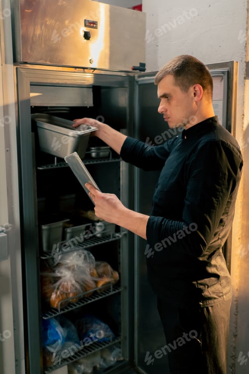 Preview: chef stands in kitchen in a black uniform and looks through what is on the list in the refrigerator