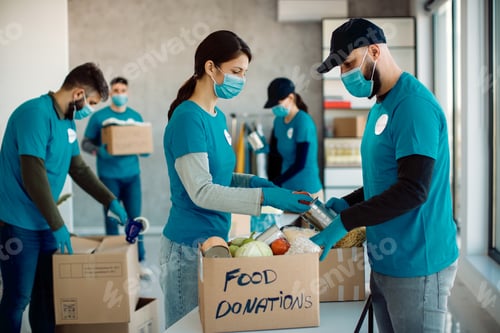 Preview: Group of volunteers with face masks packing donated food in cardboard boxes.