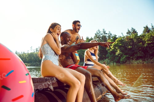 Preview: Group of happy friends having fun while sitting and laughting on the pier on river