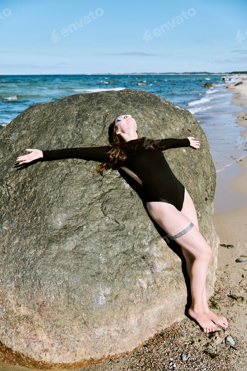 Preview: Woman relaxing against large rock on seaside beach in sunlight