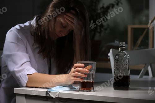 Preview: tired female doctor sitting at the table and hold glass with whiskey or cognac at home after hard