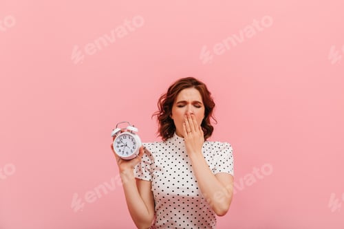 Preview: Sleepy young woman showing time. Studio shot of tired girl with clock isolated on pink background.