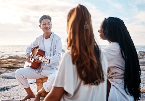 Preview: A young man at the beach playing his guitar with his two female friends. Diverse group of friends l