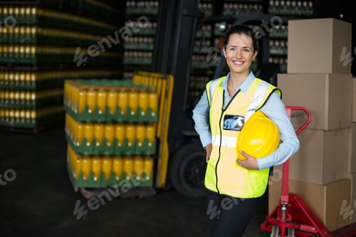 Preview: Female warehouse worker wearing safety vest holding hard hat amid beverage crates, copy space
