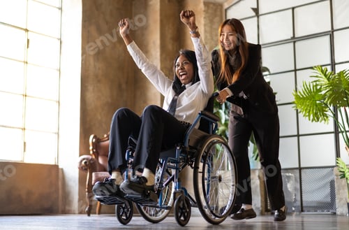 Preview: Young asian woman pushing african american collegue in wheelchair in co-working studio