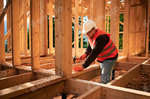 Preview: Carpenter hammering nail while constructing wooden frame two-story house near the forest.