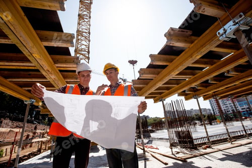 Preview: Two men dressed in shirts, orange work vests and helmets explore construction documentation on the
