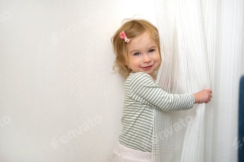 Preview: Cute caucasian blonde baby girl of 1,2 year old playing with white curtains,having fun at home