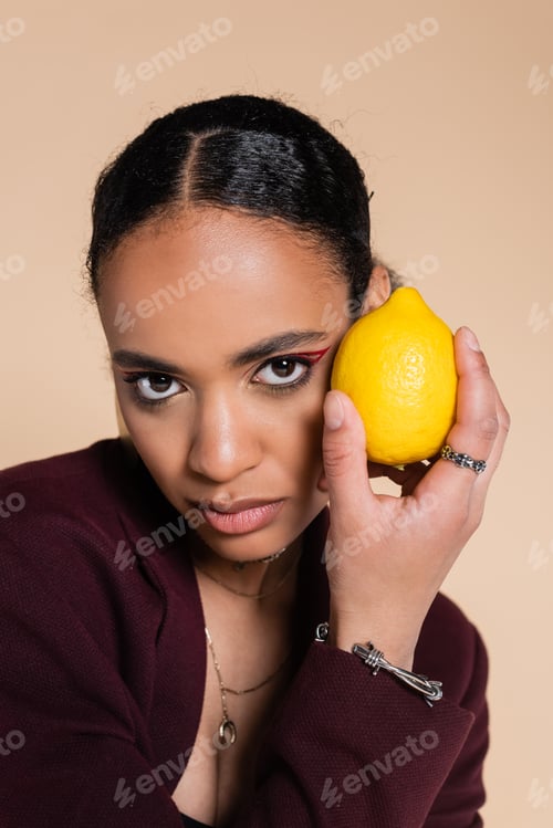 Preview: african american woman in burgundy blazer holding fresh lemon isolated on beige