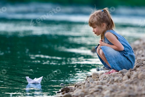 Preview: Adorable little cute blond girl in blue dress on riverbank pebbles playing with white paper origami