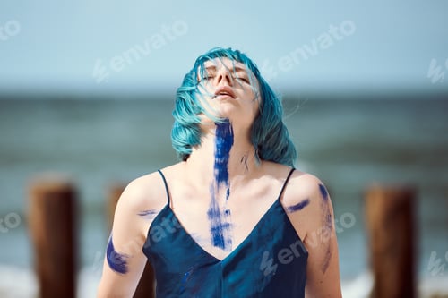 Preview: Artistic blue-haired woman performance artist smeared with blue gouache paints dancing on beach
