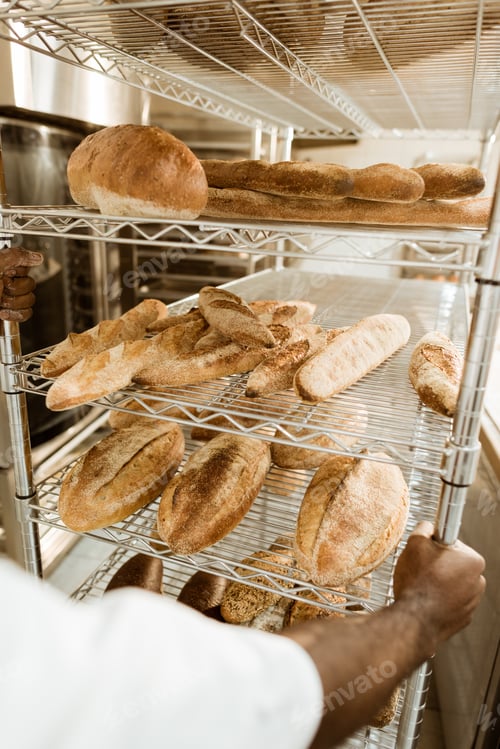 Preview: cropped shot of african american baker driving shelves of fresh bread on baking manufacture