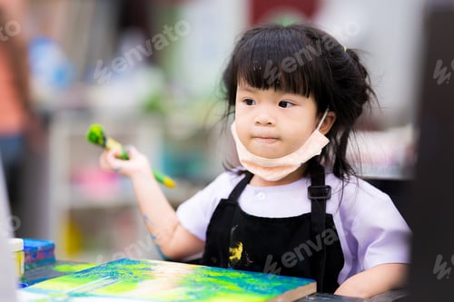 Preview: Asian little girl is sitting holding a large paintbrush and painting watercolor on a canvas.