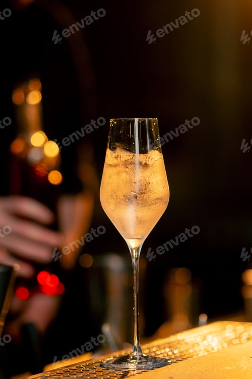 Preview: Beautiful crystal glass of bright cold cocktail and transparent shaker with ice on bar counter