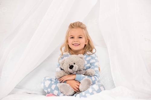 Preview: adorable child in pajamas sitting on bed and embracing teddy bear