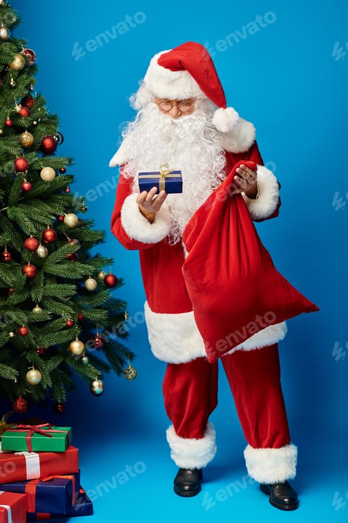 Preview: happy Santa Claus with beard and eyeglasses holding sack bag and present near Christmas tree