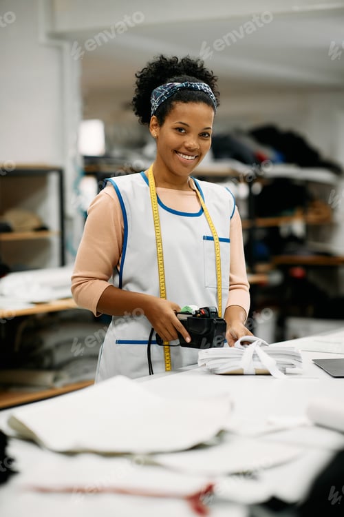 Preview: Happy black seamstress labelling finished textile products at tailor's workshop.