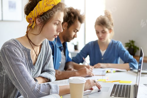 Preview: Serious female student in shirt searching for information using laptop computer drinking takeaway co