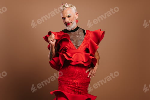 Preview: Bearded mature gay man with crown on head wearing beautiful red dress against brown background