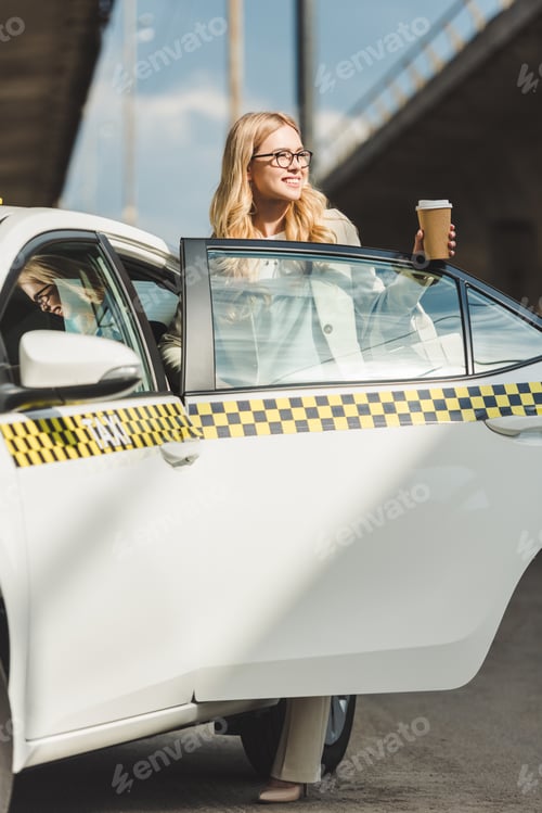 Preview: happy blonde girl in eyeglasses holding coffee to go and looking away while opening door of taxi cab