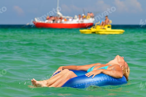 Preview: Young blond woman in blue bikini riding on swimming circle in sea and smiling