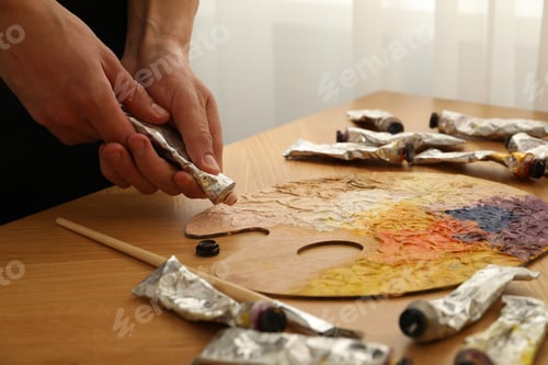 Preview: Man squeezing paint from tube onto palette at wooden table indoors, closeup