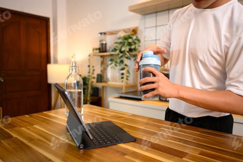 Preview: Young man checking digital tablet while preparing a protein shake on kitchen counter