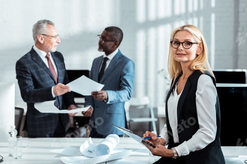 Preview: selective focus of businesswoman in glasses holding clipboard near multicultural businessmen
