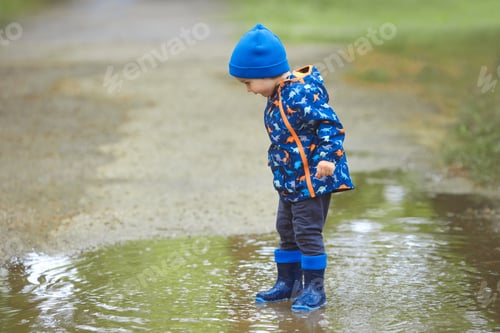 Preview: A child boy in a waterproof jacket and rubber boots runs through the puddles in the city