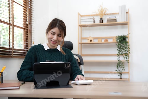 Preview: Young Asian woman using mobile phone while working at home with laptop. Female browsing site