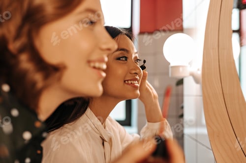 Preview: Selective focus of laughing girls doing makeup in bathroom. Indoor shot of two best friends applyin