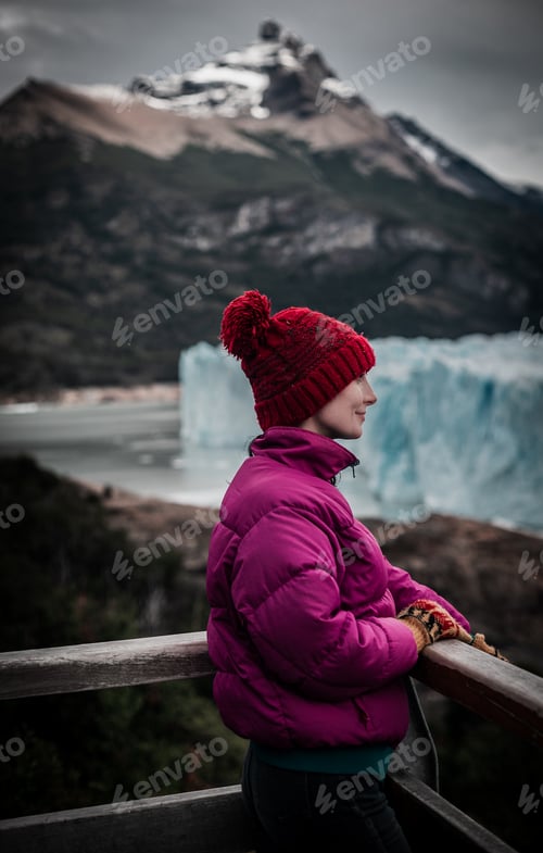 Caucasian female in a pink jacket and hat stands on a railing, admiring the majestic mountain view
