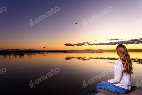 Preview: Long-haired young woman relaxing admiring the golden sunset on a beautiful pier by a natural lake.
