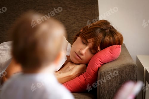 Preview: Mid adult woman sleeping on sitting room sofa watched by toddler daughter