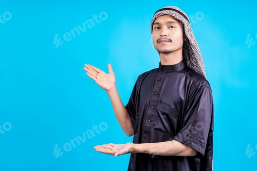 Preview: Young Arab Male Gesture Presenting To Ads Space Isolated On Blue Studio Wall