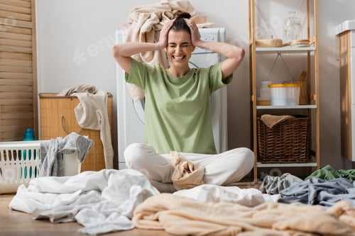 Preview: stressed young woman sitting near clothes on floor in laundry room