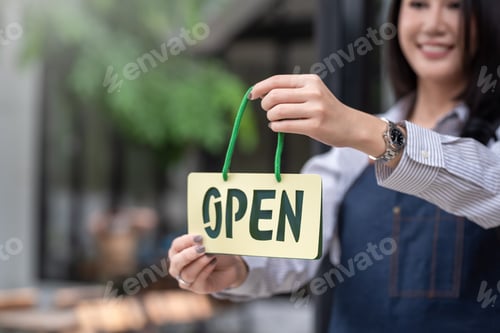 Preview: Young Asian businesswoman owner standing in front of a store holding a sign opening at the café.
