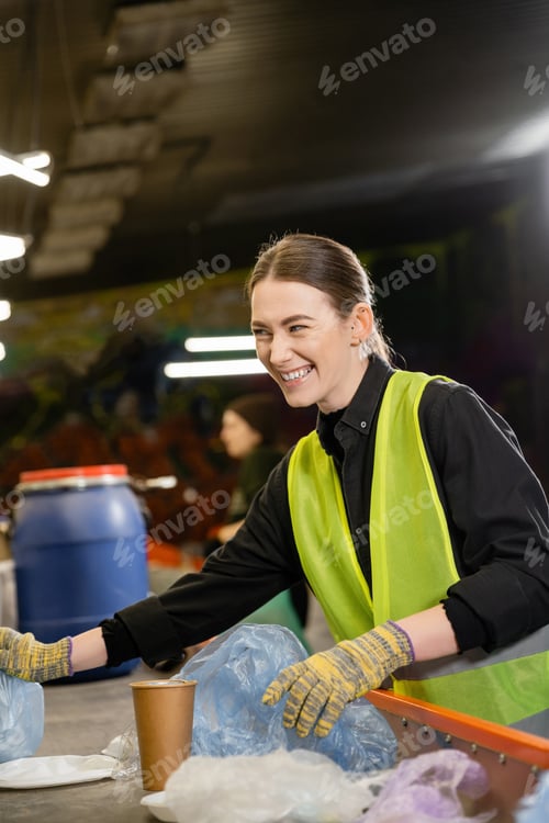 Preview: Cheerful young worker in protective vest and gloves sorting garbage on conveyor and looking away