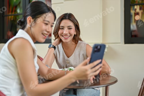 Preview: Asian woman showing photos on phone to friend sitting at wooden table in front of cafe or restaurant