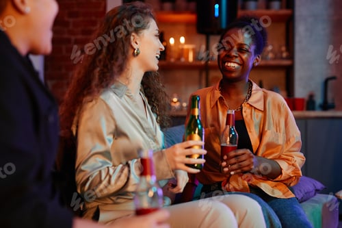 Preview: Three cheerful intercultural girls with bottles of beer having rest on couch