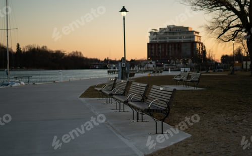 Preview: Idyllic scene of a park with a row of empty wooden benches situated along the waterfront, at dusk